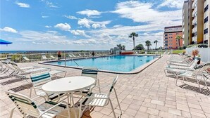 Uma área de piscina com espreguiçadeiras e uma mesa, cercada por uma grade, com vista para o oceano e um edifício ao fundo.