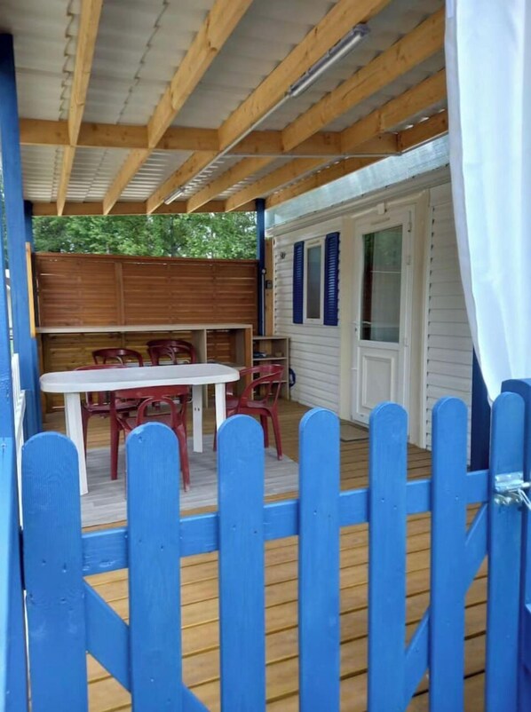 A covered outdoor area with a wooden table and chairs, a blue fence, and a white door with blue shutters. - LAC du DER , Mobilhome 4 Personnes (Châtillon-sur-Broué)
