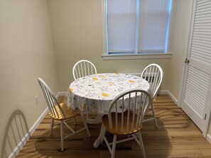 A dining area with a round table covered by a floral tablecloth, surrounded by four white chairs with wooden seats.
