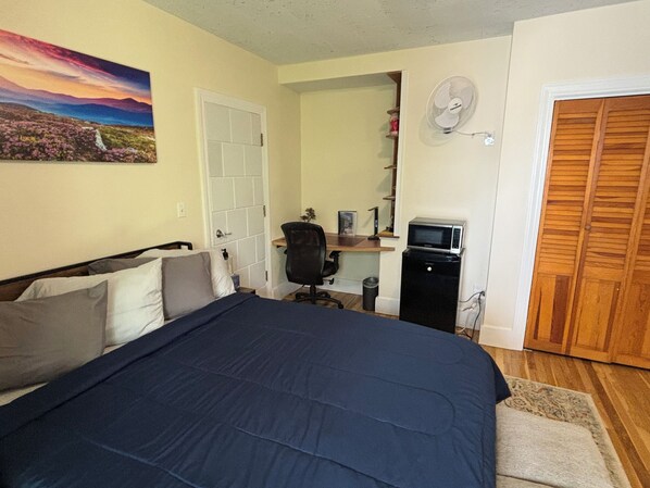 A bedroom with a bed, a desk, a chair, a microwave, a fan, and a door.