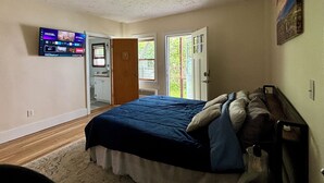 A bedroom with a bed, a television mounted on the wall, a door leading to a bathroom, and a window with a view of greenery outside. - Traveler’s Den in Medford (Medford)