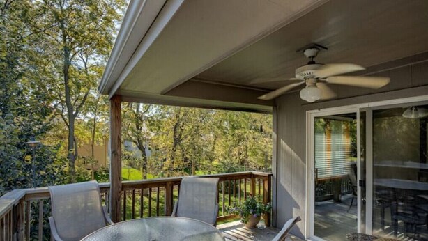 A covered outdoor area with a glass table and chairs, a ceiling fan, and a view of a garden.