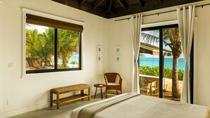Een slaapkamer met een groot bed, een bankje, een stoel en een kleine tafel. Er is een raam met uitzicht op palmbomen en een strand.