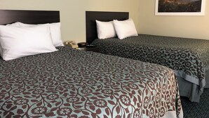 Ein Hotelzimmer mit zwei Betten, jeweils mit gemusterten Bettbezügen und weißen Kissen. Ein Nachttisch mit Telefon und Fernbedienung ist vorhanden. An der Wand hängt ein gerahmtes Bild eines Sonnenuntergangs.