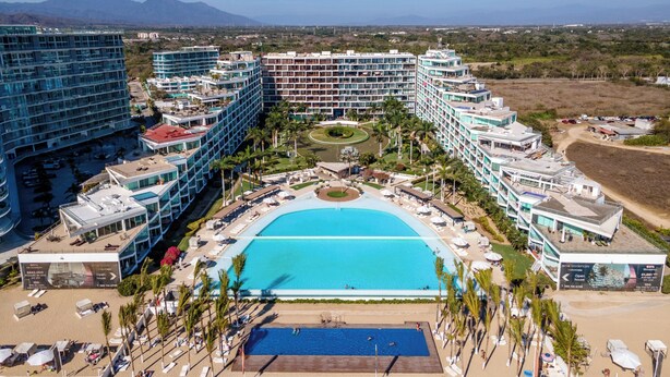 Un hotel grande con varias edificaciones, una piscina central y una zona de playa con tumbonas y sombrillas.
