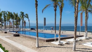 Zona de piscina con vista despejada al océano, rodeada de palmeras y sillones de descanso.