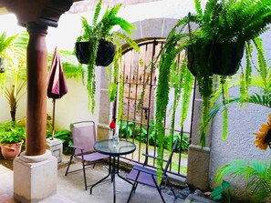 Un espace patio avec une table et des chaises en verre, entouré de plantes en pot et d’un portail en fer forgé.