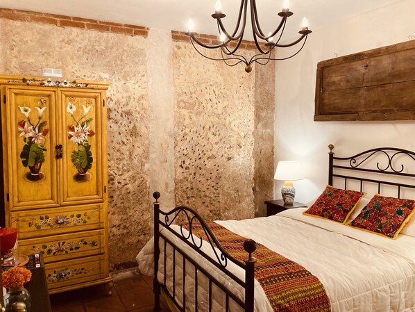 A bedroom with a bed, a wooden wardrobe with floral designs, a chandelier, and a nightstand with a lamp. - Luna Maya Antigua Hotel (Antigua Guatemala)
