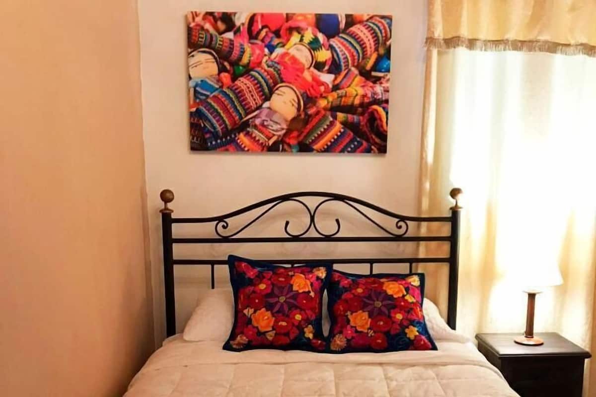 A bed with a quilt and two floral pillows, a metal headboard, a small bedside table with a lamp, and a colorful artwork on the wall.