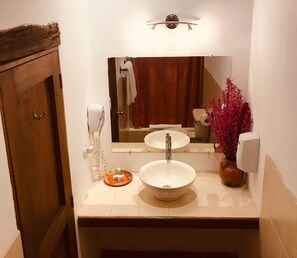 A bathroom with a wooden door, a white sink, a mirror, a towel rack, a hand dryer, and a decorative plant. - Luna Maya Antigua Hotel (Antigua Guatemala)
