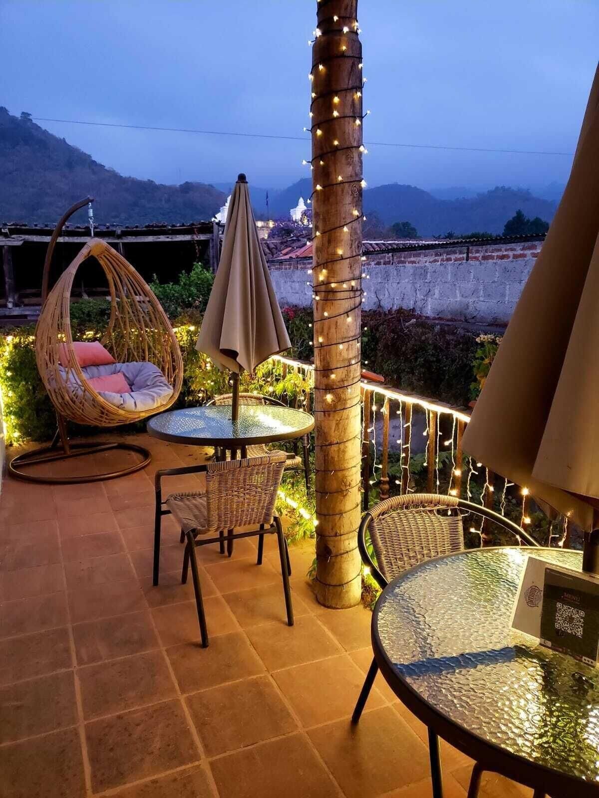 Área externa com mobiliário de vime, uma mesa redonda e um pilar decorativo com iluminação.