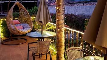 Área externa com mobiliário de vime, uma mesa redonda e um pilar decorativo com iluminação.