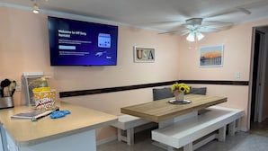 A dining area with a wooden table, benches, a TV displaying a welcome message, a popcorn container, and a wall with a 'USA' sign and a sunset painting. - Off Duty Retreat ~Sleeps 10, Boat Slip, Screen Porch, Lots of Parking, 2 Grills (Steinhatchee)