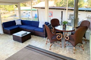 Un patio couvert comprenant un canapé d’angle bleu, des fauteuils en osier, une table avec un plateau en verre et une petite plante en pot.
