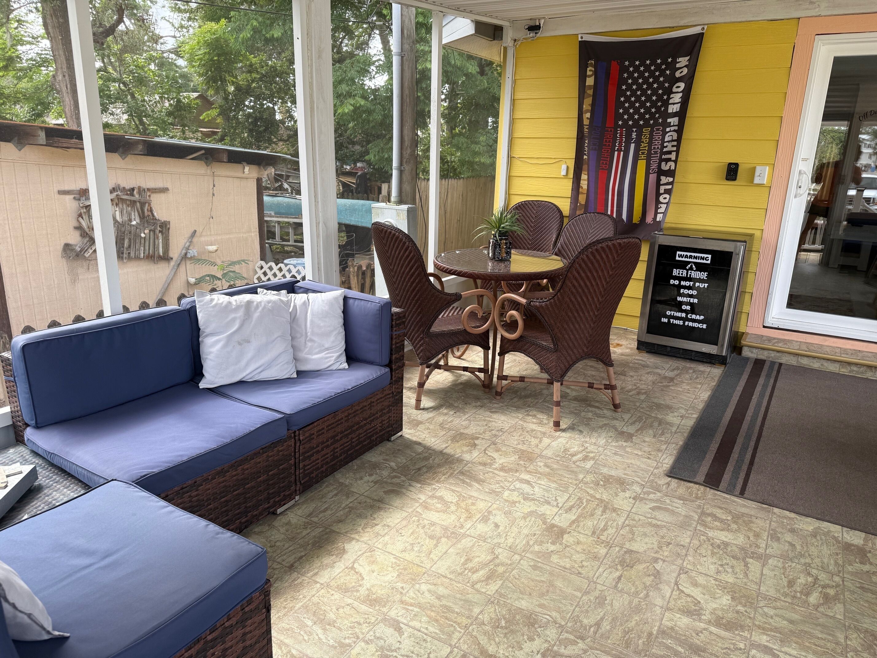 Un espace patio avec un canapé bleu, des chaises en osier et une petite table. Un mur jaune est décoré de drapeaux et porte un panneau d’avertissement interdisant de mettre de la nourriture ou de l’eau dans le réfrigérateur.