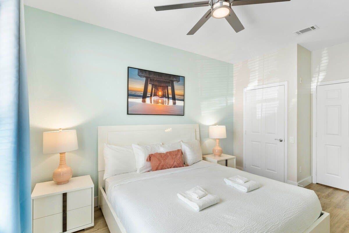 Une chambre d’hôtel avec un lit, deux tables de chevet, un ventilateur de plafond, un tableau encadré au mur et une porte blanche.