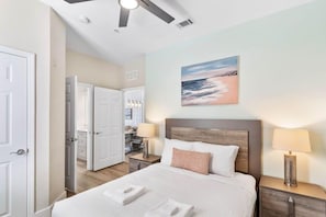 A hotel room with a bed, bedside lamps, a painting of a beach scene, and a ceiling fan.