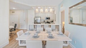 Un espace repas moderne avec une table blanche dressée pour six personnes, une cuisine en arrière-plan et un grand miroir accroché au mur.