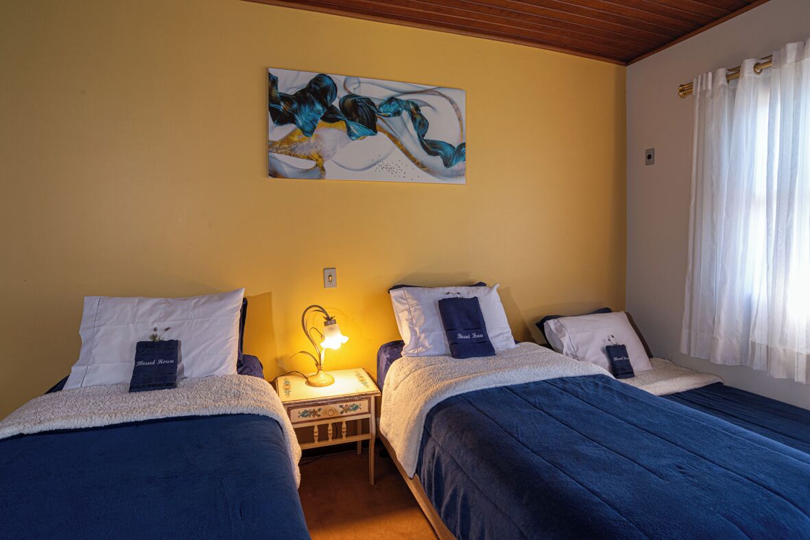 A room with two beds, a nightstand with a lamp, a painting on the wall, and a window with curtains.