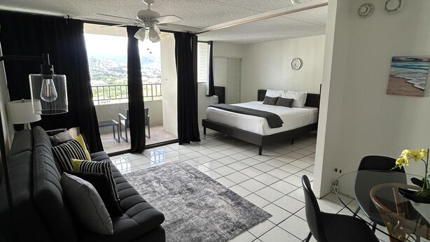 A modern hotel room with a bed, a sofa, a dining table, and a balcony with curtains. - Heart of Waikiki w/ Kitchen - Free Parking - King Bed - Mountain View (Honolulu)