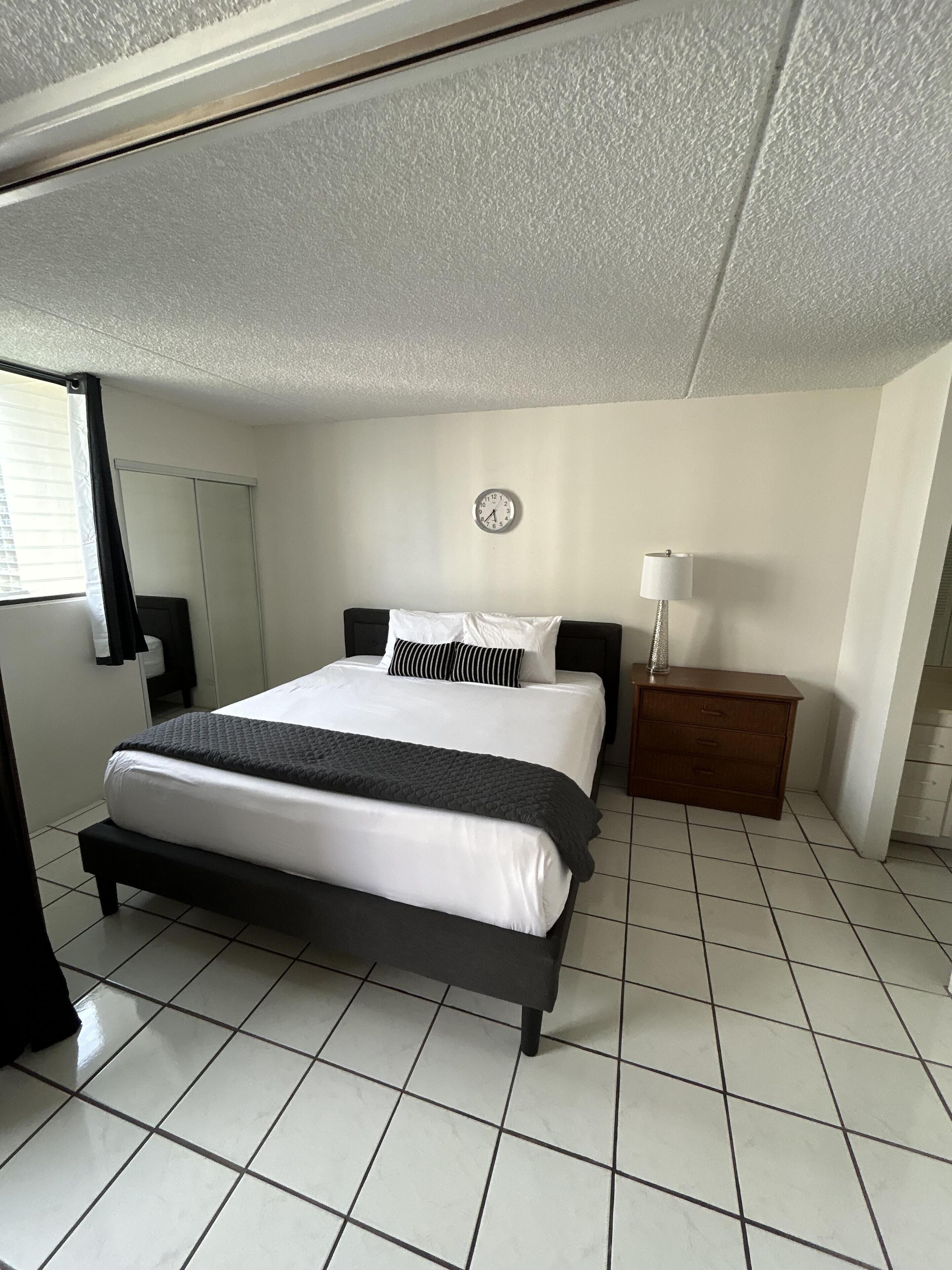 Quarto com cama, mesa de cabeceira, abajur, relógio e janela com cortinas.