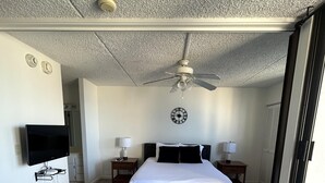 A modern bedroom with a bed, two bedside tables, a ceiling fan, a TV mounted on the wall, and a clock.