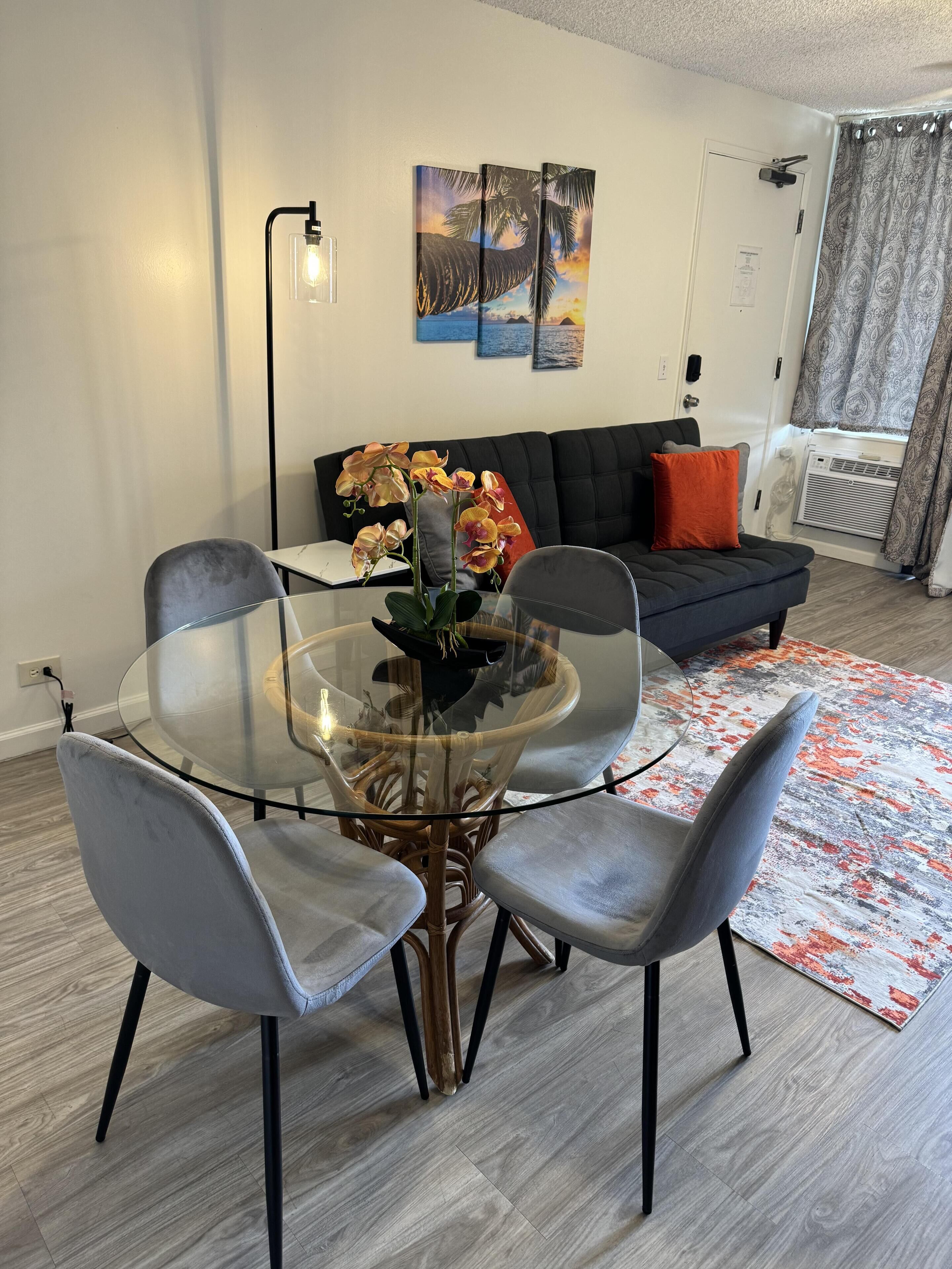 En moderne stue med et glasbord omgivet af stole, en sort sofa med orange puder og en tæppe med et mønster.