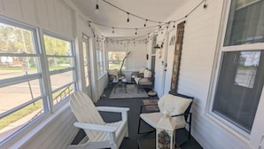 A covered porch with white chairs, a small table, and string lights. - Downtown Muskegon Hideaway (Muskegon)