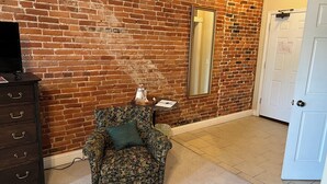 Une pièce avec un mur en briques, une commode en bois, un fauteuil à motifs floraux, un miroir et une porte blanche.