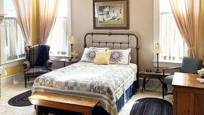 Une chambre à coucher avec un lit, deux chaises, un bureau, un ventilateur de plafond et un tableau encadré accroché au mur.