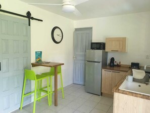 Une cuisine compacte équipée d’un réfrigérateur, d’un four à micro-ondes et de meubles de rangement en bois. Une petite table avec deux chaises est placée près de la porte.