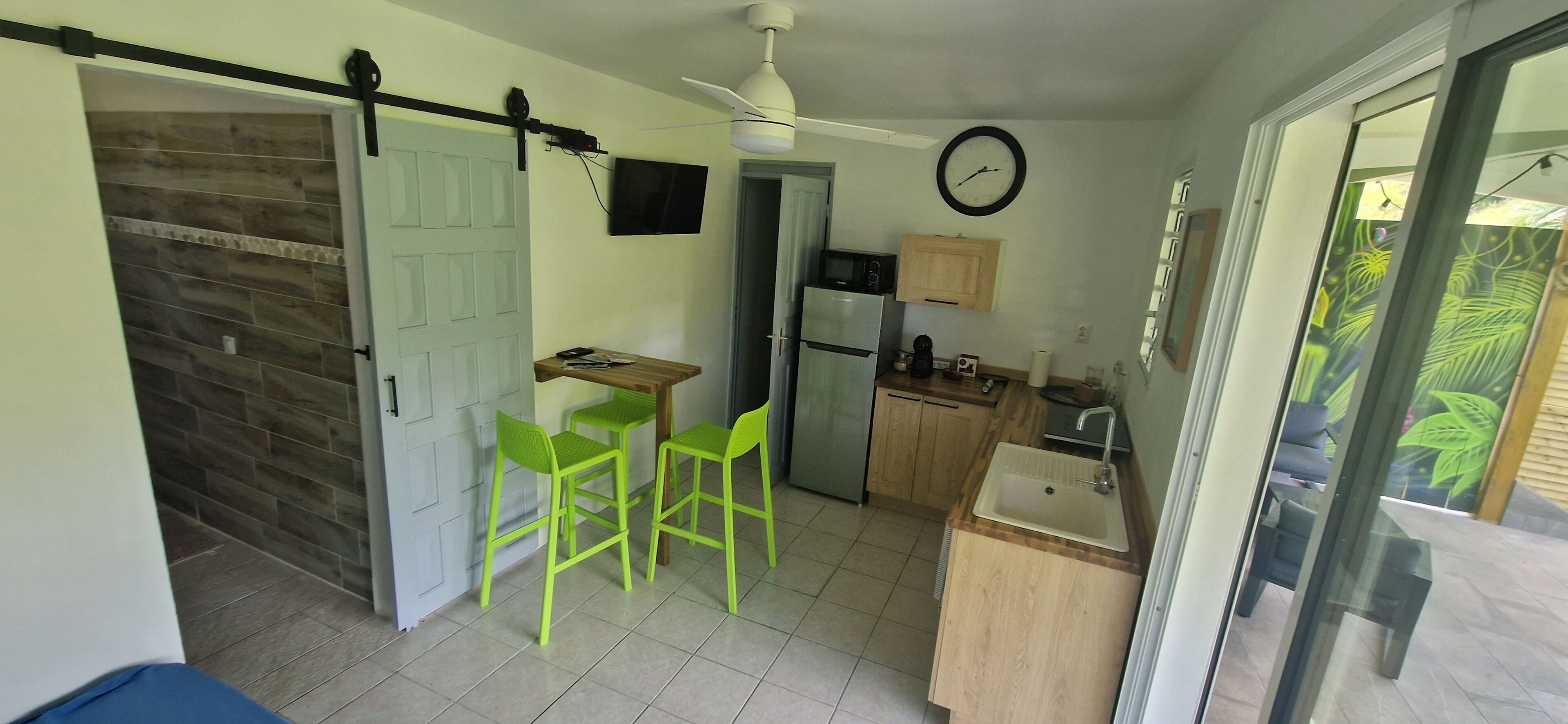 Une cuisine compacte équipée d’un four à micro-ondes, d’un réfrigérateur et de meubles en bois. On y trouve deux chaises vertes et une petite table. Une horloge est fixée au mur.