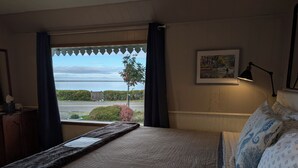 Un dormitorio con cama, ventana con cortinas, un cuadro en la pared y vista al mar.