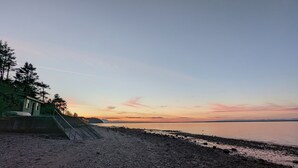Una playa con una estructura de madera, una escalera que baja hacia la orilla y un mar tranquilo bajo un cielo al atardecer.