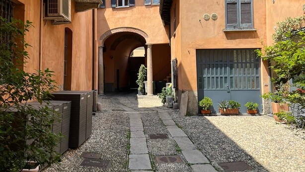 A courtyard with a stone pathway, an archway, and buildings with balconies and shutters. - Fashionable by the canals (Milano)