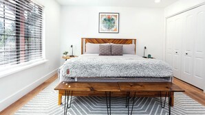 Una camera da letto moderna con una testiera in legno, una coperta grigia e soffice, una panca ai piedi del letto, un comodino con una lampada e un'ampia finestra con persiane.