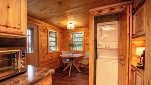 Interior de una cabaña de madera con zona de cocina que incluye un microondas, una mesa de comedor con sillas y una lavadora.