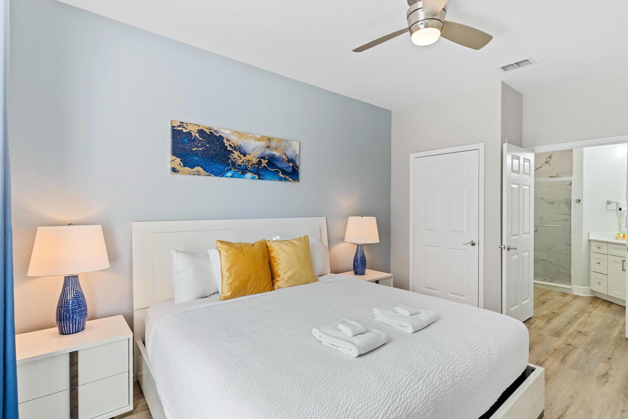 Une chambre à coucher bien rangée, avec un lit, deux tables de chevet dotées de lampes, un ventilateur de plafond, une porte et une salle de bain visible à travers une porte ouverte.