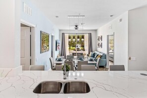 A modern kitchen with a double sink, dining area with a table and chairs, and a living room with a sofa and a view of the outdoors.