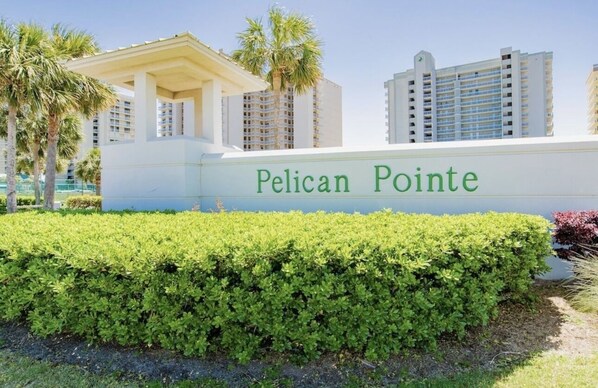 A sign for Pelican Pointe with a white building and a well-maintained hedge in front. - 5 STARS Beachfront Condo, Indoor/Outdoor Pools, Hot Tub & Tennis! BOOK NOW! (Orange Beach)