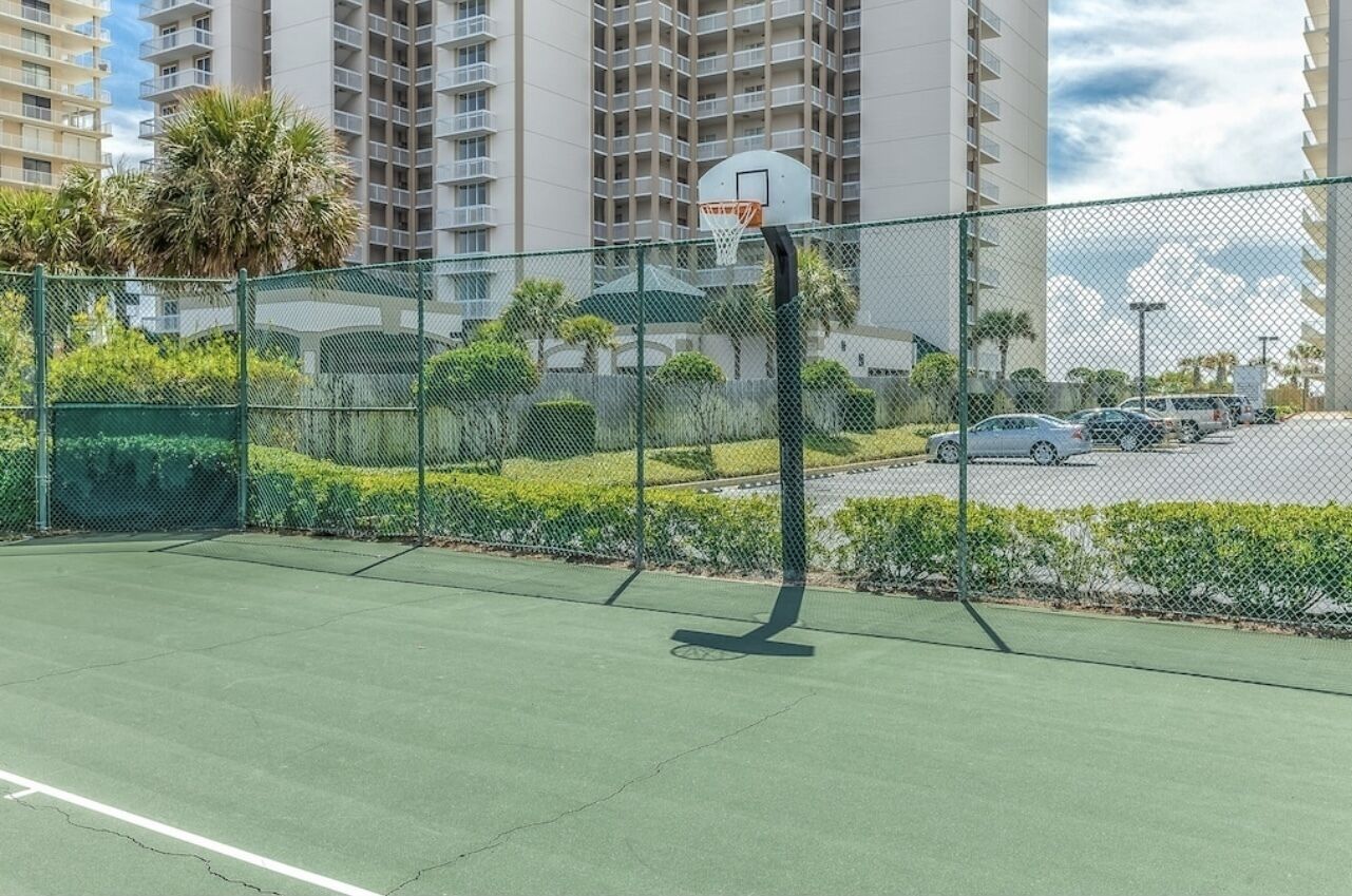 A basketball court with a chain-link fence, surrounded by greenery and tall buildings in the background.