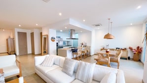 A modern living room with a white sofa, dining area with wicker chairs, and a kitchen in the background.