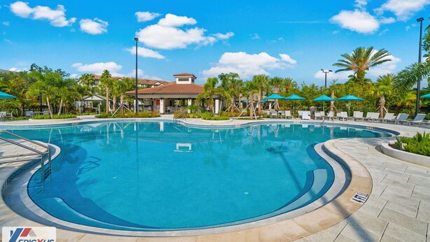 A swimming pool with a curved edge, surrounded by lounge chairs and umbrellas, and a building with a red-tiled roof in the background.