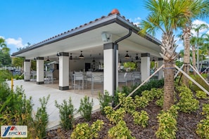 A covered outdoor seating area with tables and chairs, surrounded by plants and palm trees.