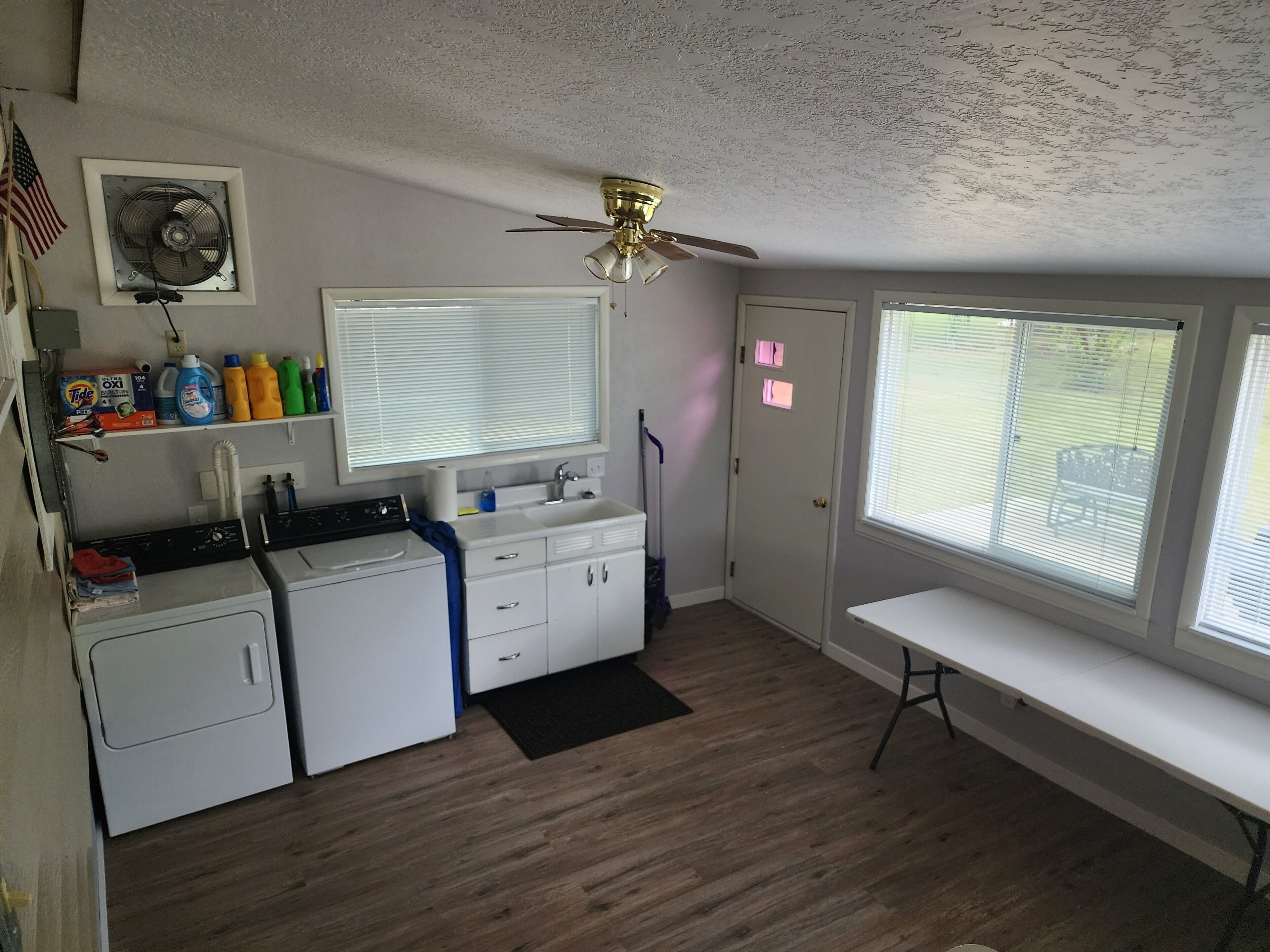 Uma lavanderia com máquina de lavar, secadora e ventilador no teto. Há uma janela com persianas e uma porta com uma placa rosa.