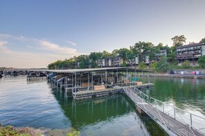 En brygga vid vattnet med ett överbyggt sittområde, en bro och byggnader i bakgrunden.
