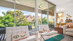A modern living room with a large glass door leading to a balcony, white sofas with decorative pillows, a small table, and a chandelier. - Lakeside Luxe: Seven Lakes Golf & Country Club stylish condo (Palm Springs)