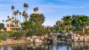 A landscaped garden with a pond, palm trees, and a building with a flat roof. - Lakeside Luxe: Seven Lakes Golf & Country Club stylish condo (Palm Springs)