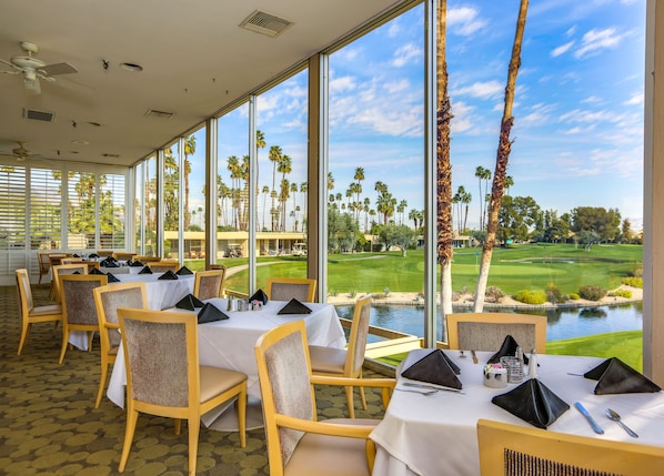 A dining area with tables set for a meal, large windows offering a view of a golf course and palm trees.