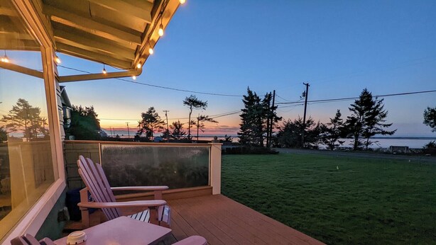 En træterrasse med et bord og stole, der vender ud mod et græsareal og et vandareal i solnedgang.
